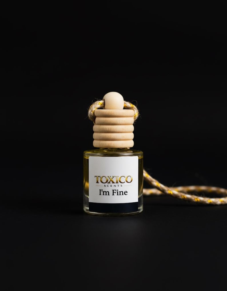 Puede incluir: Una peque&ntilde;a botella de vidrio transparente de ambientador Toxico Scents con el texto "I'm Fine" en negro y dorado. La botella tiene una tapa de madera y una cuerda decorativa. El fondo es negro.