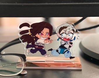 May include: A clear acrylic standee featuring two cartoon characters in a running pose. One character has long dark brown hair and a wide grin, while the other has short white hair, a panicked expression, and holds a blue and white curved object.