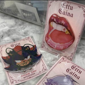 May include: Several phone grips are displayed, each packaged in clear plastic. One features a design of red lips with fangs, another depicts a character with red and blue accents, and the third has a purple and white design. Each grip has the text "Ettu Caina".