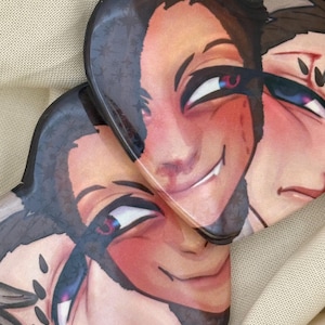 May include: Two heart-shaped pins featuring a stylized illustration of a character with dark hair, a small fang, and flushed cheeks. The artwork includes a subtle star pattern overlay on the dark hair and expressive eyes with pink and blue highlights.