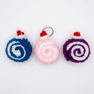 Könnte beinhalten: Drei handgefertigte Häkelornamente in Blau, Rosa und Lila mit weißen Spiralmustern. Jedes Ornament hat einen kleinen roten gehäkelten Hut und einen Metall-Schlüsselring. Auf weißem Hintergrund.