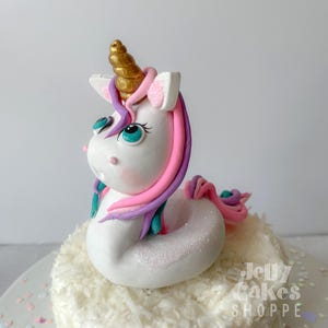 May include: A white unicorn cake topper with a golden horn, pink, purple, and teal accents. The unicorn has large blue eyes and is sitting on a white cake with a coconut topping. The cake is decorated with iridescent sprinkles.