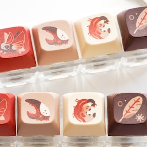 Peut inclure: Un ensemble de six touches de clavier avec des motifs différents. Les touches sont de couleur rouge, marron et beige. Chaque touche présente une illustration différente, notamment un papillon, une griffe, un oiseau et une feuille. Les touches sont attachées à un porte-clés en plastique transparent.
