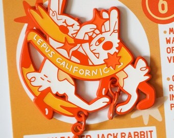 Könnte beinhalten: Orangefarbener und weißer Emaille-Pin mit einem Cartoon-Schwarzschwanz-Kaninchen mit dem Text "LEPUS CALIFORNIC" und "BLACK-TAILED JACK RABBIT".