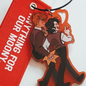 May include: A red fabric patch with white embroidery that reads "FOR ANYTHING FOR OUR MOONY."  A clear acrylic charm with a cartoon illustration of two men holding hands and smiling. The man on the left is wearing a brown suit and the man on the right is wearing a brown vest and black pants.