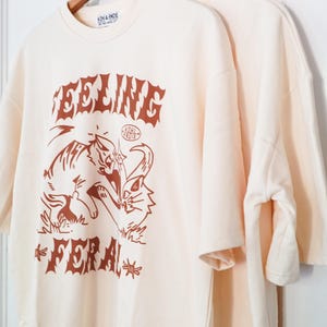 K&ouml;nnte beinhalten: Cremefarbene T-Shirts mit braunem Grafikdesign. Das Design zeigt das Wort "FEELING" &uuml;ber einer Tiger- und Katzenillustration, darunter das Wort "FERAL". Der Markenname "KOHL & ANDIE" ist oben aufgedruckt.