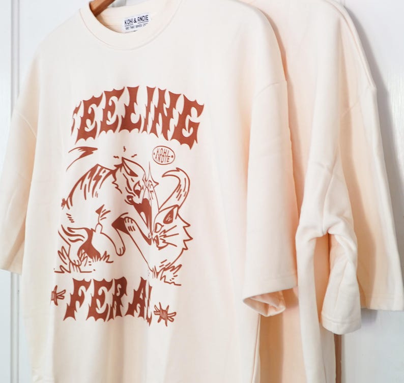 K&ouml;nnte beinhalten: Cremefarbene T-Shirts mit braunem Grafikdesign. Das Design zeigt das Wort "FEELING" &uuml;ber einer Tiger- und Katzenillustration, darunter das Wort "FERAL". Der Markenname "KOHL & ANDIE" ist oben aufgedruckt.