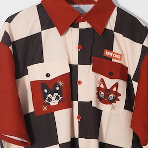 Peut inclure: Chemise à manches courtes à carreaux noirs, crème et rouges. Elle comporte deux poches avant avec des broderies de visages de chats et le texte "SAVE CATS".