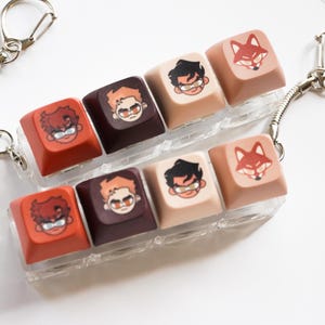 May include: Keycaps with cartoon character designs in red, brown, and tan. Each keycap features a different character or a fox illustration. The keycaps are attached to a clear plastic holder with a silver chain.