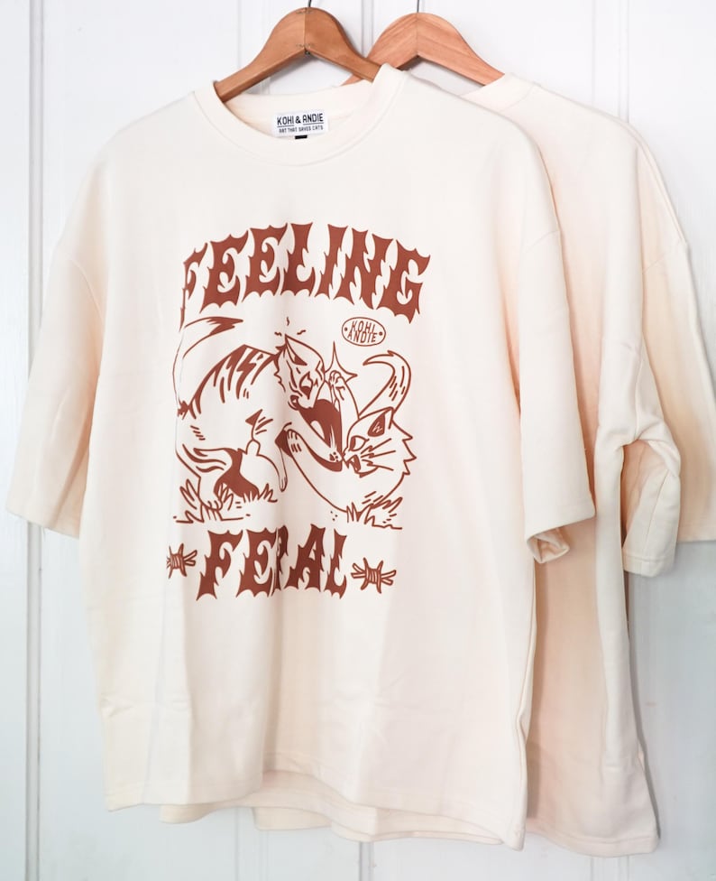 K&ouml;nnte beinhalten: Zwei cremefarbene T-Shirts h&auml;ngen an Holzb&uuml;geln. Das vordere Shirt zeigt eine braune Grafik von zwei Katzen und die Worte "FEELING FERAL" in einer stilisierten Schriftart. Der Markenname "KOHI & ANDIE" ist oben aufgedruckt.