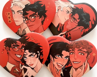 May include: Four heart-shaped badges featuring illustrations of male characters. The badges have a red background with illustrations in brown, black, and white. Each badge depicts two characters in various poses, with a fantasy theme.