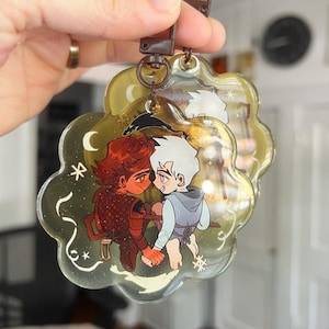 Può includere: Un portachiavi trasparente a forma di nuvola con un'illustrazione a cartoni animati di due figure che si tengono per mano. Una figura ha i capelli rossi e un'armatura, l'altra ha i capelli bianchi e una felpa con cappuccio blu. Il portachiavi ha un cinturino marrone e accessori in metallo.
