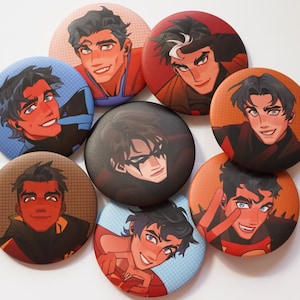 Puede incluir: Una colección de ocho botones redondos con ilustraciones estilizadas de personajes masculinos. Los botones muestran varios esquemas de color, incluyendo rojo, azul y naranja, con expresiones faciales detalladas y diseños de personajes. Los botones están dispuestos sobre una superficie blanca.