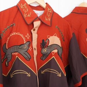 May include: Rust-colored button-down shirt with embroidered collar and animal graphics. Features a wolf and rabbit design with sun and moon accents. The shirt has brown sleeves and a tan front panel.