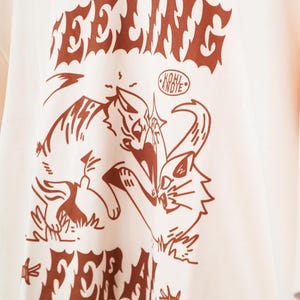 K&ouml;nnte beinhalten: Cremefarbenes T-Shirt mit einer braunen Grafik von zwei k&auml;mpfenden Katzen, mit den Worten "FEELING" und "FERAL" &uuml;ber und unter dem Bild. Der Text "KOHL ANDIE" befindet sich in einem Kreis &uuml;ber den Katzen. Das Shirt hat kurze &Auml;rmel.
