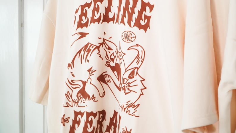 K&ouml;nnte beinhalten: Cremefarbenes T-Shirt mit einer braunen Grafik von zwei k&auml;mpfenden Katzen, mit den Worten "FEELING" und "FERAL" &uuml;ber und unter dem Bild. Der Text "KOHL ANDIE" befindet sich in einem Kreis &uuml;ber den Katzen. Das Shirt hat kurze &Auml;rmel.