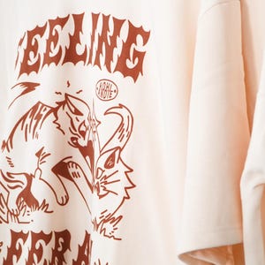 K&ouml;nnte beinhalten: Cremefarbenes T-Shirt mit braunem Grafikdesign. Das Design zeigt das Wort "FEELING" in einer stilisierten Schriftart &uuml;ber einer Zeichnung einer katzenartigen Kreatur. Das Shirt hat kurze &Auml;rmel und eine lockere Passform.