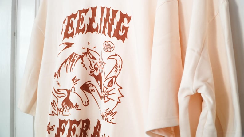 K&ouml;nnte beinhalten: Cremefarbenes T-Shirt mit braunem Grafikdesign. Das Design zeigt das Wort "FEELING" in einer stilisierten Schriftart &uuml;ber einer Zeichnung einer katzenartigen Kreatur. Das Shirt hat kurze &Auml;rmel und eine lockere Passform.