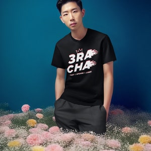 May include: A man wearing a black t-shirt with a white graphic that says "3RACHA" with a crown and flames. The text below the graphic says "CB97 | SPEARB | J.ONE". He is standing in a field of white and pink flowers.