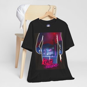 May include: Black t-shirt with a graphic design featuring a building scene with the text "Killer Frequency" in pink and purple. A white baseball cap hangs on a wooden chair in the background.
