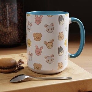 Puede incluir: Una taza de cer&aacute;mica blanca con un borde y un asa azules. La taza presenta un patr&oacute;n repetido de caras de animales de dibujos animados, incluidos conejos, gatos, perros y p&aacute;jaros. La taza est&aacute; sobre una superficie de madera con una cuchara y un macaron.