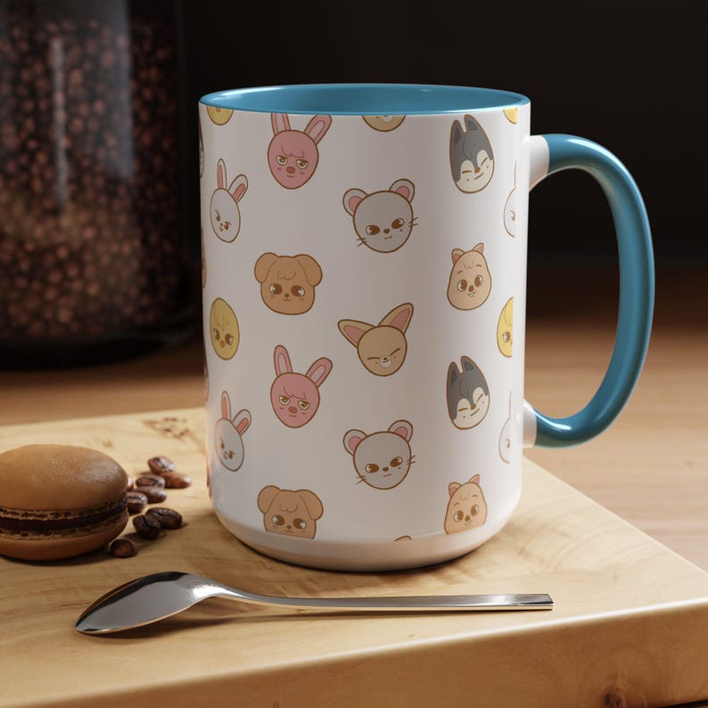Puede incluir: Una taza de cer&aacute;mica blanca con un borde y un asa azules. La taza presenta un patr&oacute;n repetido de caras de animales de dibujos animados, incluidos conejos, gatos, perros y p&aacute;jaros. La taza est&aacute; sobre una superficie de madera con una cuchara y un macaron.