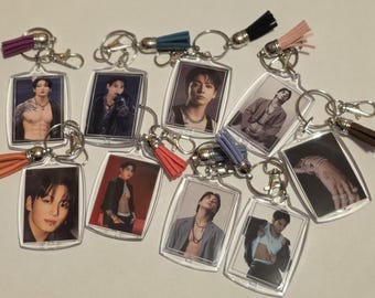 May include: A collection of nine rectangular acrylic keychains featuring various photographic portraits of a person with dark hair. Each keychain includes a metal ring, a lobster clasp, and a small decorative tassel in colors including purple, blue, black, pink, orange, and brown.