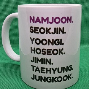 Pu&ograve; includere: Una tazza in ceramica bianca con un elenco verticale di nomi stampati in testo nero e viola. I nomi elencati sono NAMJOON, SEOKJIN, YOONGI, HOSEOK, JIMIN, TAEHYUNG e JUNGKOOK. La tazza presenta un manico curvo sul lato sinistro e finitura liscia.