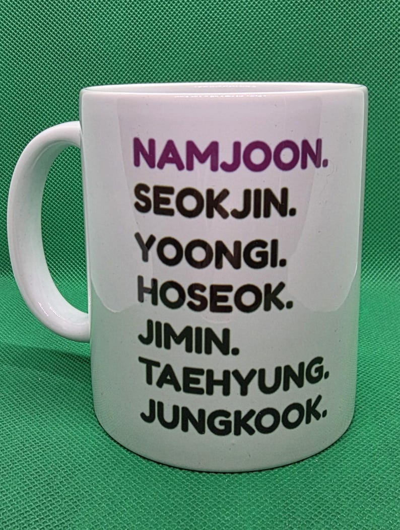 Pu&ograve; includere: Una tazza in ceramica bianca con un elenco verticale di nomi stampati in testo nero e viola. I nomi elencati sono NAMJOON, SEOKJIN, YOONGI, HOSEOK, JIMIN, TAEHYUNG e JUNGKOOK. La tazza presenta un manico curvo sul lato sinistro e finitura liscia.