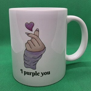 Pu&ograve; includere: Una tazza in ceramica bianca con un'illustrazione grafica di una mano che fa il gesto del cuore con le dita e un piccolo cuore viola sopra. Il testo "I purple you" &egrave; stampato in caratteri neri sotto l'illustrazione sulla parte anteriore.