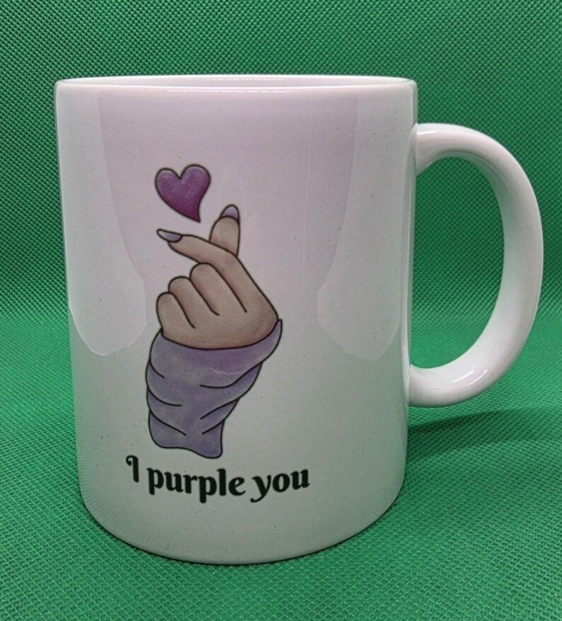 Pu&ograve; includere: Una tazza in ceramica bianca con un'illustrazione grafica di una mano che fa il gesto del cuore con le dita e un piccolo cuore viola sopra. Il testo "I purple you" &egrave; stampato in caratteri neri sotto l'illustrazione sulla parte anteriore.