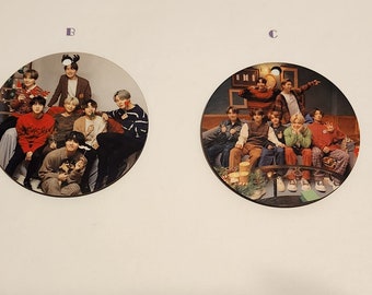 Op de afbeelding: Een set van vijf ronde, houten ornamenten met afbeeldingen van een populaire K-pop-groep. De voorkant van de ornamenten toont verschillende afbeeldingen van de groep, terwijl de achterkant de naam van de groep, "BTS", in witte letters op een rode achtergrond toont.