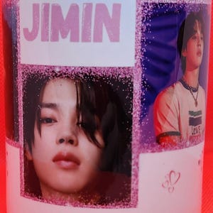 May include: White ceramic mug with a pink glitter design. The mug features a collage of photos of a young man with the word "JIMIN" printed in pink letters.