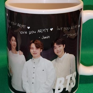 May include: White ceramic mug with a black background featuring a photo of four men. The mug has the text "love you! ARMY" and "love you ARMY" written on it. The text "BTS" is written in large white letters on the bottom of the mug.