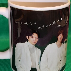 May include: White ceramic mug with a black background featuring a photo of two men in white clothing. The text "love you ARMY" is written on the mug in white, with the names "V" and "Jin" written below.