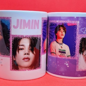 May include: Two white ceramic mugs with a purple and pink design featuring a Korean pop star. The mug on the left has the word "JIMIN" printed on it. The mug on the right has the word "LOVE" printed on it.