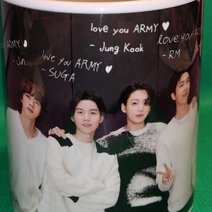 May include: A white ceramic mug with a black background featuring a photo of four men. The mug has handwritten messages in white chalk that say "love you ARMY" and "love you ARMY" with the names "Jung Kook", "RM", "Jin", and "SUGA" written below.