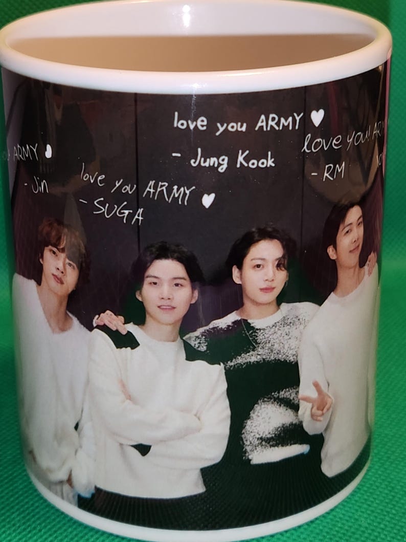 May include: A white ceramic mug with a black background featuring a photo of four men. The mug has handwritten messages in white chalk that say "love you ARMY" and "love you ARMY" with the names "Jung Kook", "RM", "Jin", and "SUGA" written below.