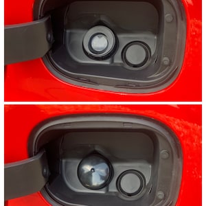 May include: Close-up of a red car's fuel door with two round openings. The top opening has a black cap, while the bottom opening has a silver cap.