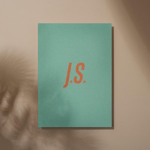 Könnte beinhalten: Eine türkisgrüne Karte mit den orangefarbenen Buchstaben "J.S." und zwei kleinen orangefarbenen Sternen. Die rechteckige Karte hat eine strukturierte Oberfläche. Der Hintergrund ist beige.
