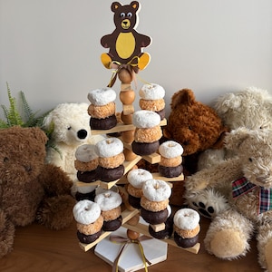 May include: A wooden tiered donut display stand featuring a cutout bear topper, a central pole with spherical accents, and horizontal arms holding stacks of white powdered, toasted coconut, and dark chocolate donuts on a hexagonal base.