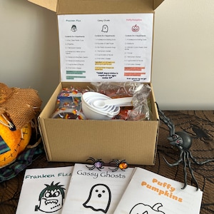 May include: A Halloween-themed science kit in a cardboard box, with instructions for experiments like "Franken Fizz," "Gassy Ghosts," and "Puffy Pumpkins." The box contains measuring cups, ingredients, and log books. A decorative spider and pumpkin are in the background.