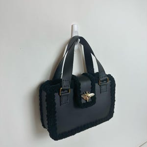 Może przedstawiać: Czarna torebka o prostokątnym kształcie z górnym uchwytem. Torebka posiada złoty emblemat pszczoły na przedniej klapie i jest obszyta ciemnym, puszystym materiałem. Torebka wisi na białej ścianie.