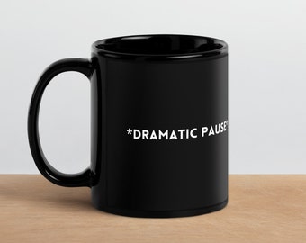 May include: A black ceramic coffee mug with a white inscription that reads "*DRAMATIC PAUSE*" on a wooden surface.