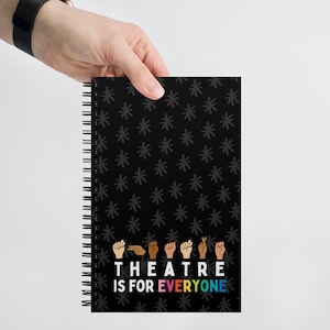 Pode incluir: Caderno preto de espiral com um padrão de estrelas preto e branco. O caderno tem o texto "THEATRE IS FOR EVERYONE" em cores do arco-íris com uma fileira de mãos em diferentes tons de pele fazendo a linguagem de sinais para a palavra "teatro".