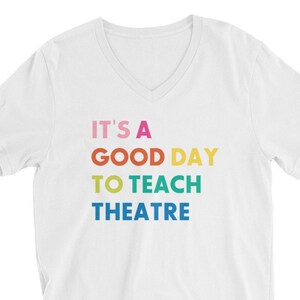 May include: White V-neck t-shirt with the text "It's a good day to teach theatre" in rainbow colours.