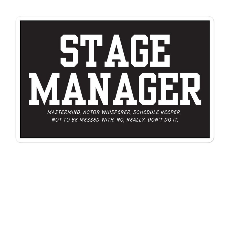 May include: Black and white sticker with the text "STAGE MANAGER" in large white letters. Below the text is a smaller line of text that reads "MASTERMIND, ACTOR WHISPERER, SCHEDULE KEEPER. NOT TO BE MESSED WITH, NO, REALLY, DON'T DO IT."