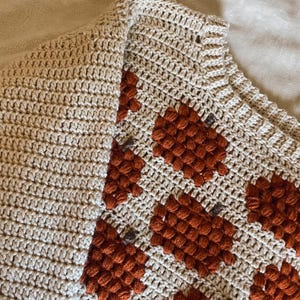 Może przedstawiać: Beżowy sweter dziergany na szydełku z brązowym i pomarańczowym geometrycznym wzorem. Sweter ma fakturowaną, nierówną powierzchnię.