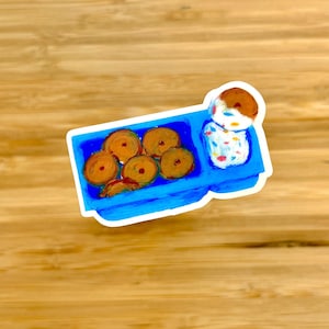 May include: A sticker featuring a blue lunch tray with a variety of food items. The tray contains several round, golden-brown pastries and a small dessert with a white base and a brown top. The sticker is on a wooden surface.