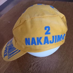 May include: A yellow cycling cap featuring the blue text NAKAJIMA and the number 2 printed on the side. The brim of the cap displays the blue text FORMULA 1 GRAND PRIX. The cap is constructed from a durable fabric with a structured, multi-panel design.